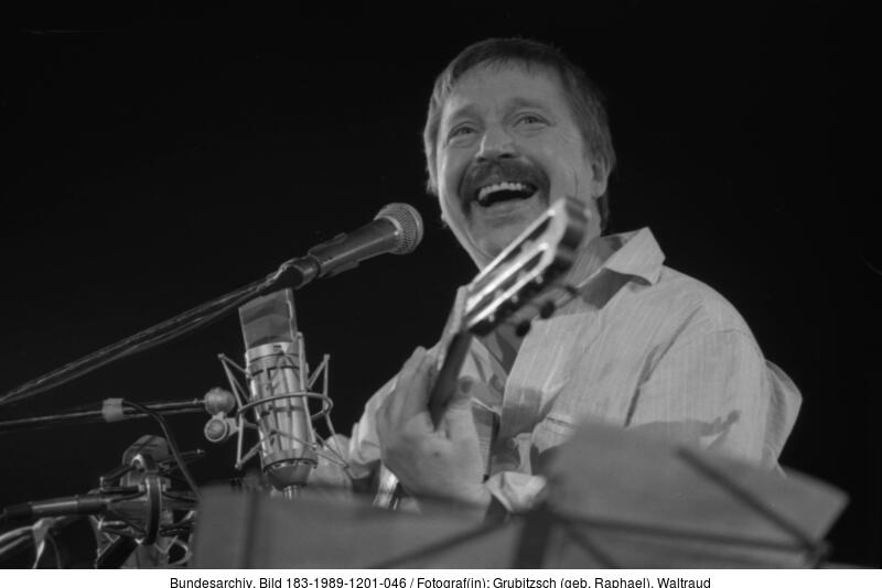 Schwarz-Weiß-Foto eines lächelnden, Gitarre spielenden Mannes, der auf der Bühne in ein Mikrofon singt.