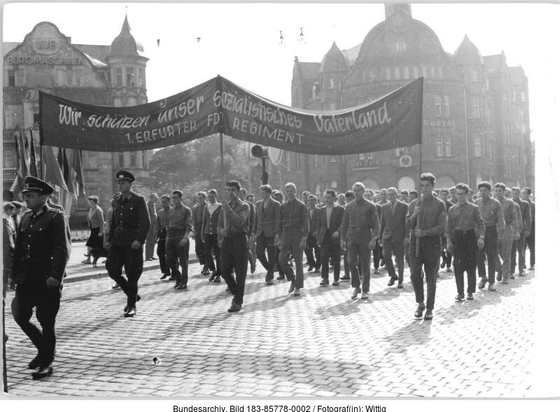 Eine Gruppe uniformierter Männer marschiert in Formation auf einer Kopfsteinpflasterstraße und trägt eine große Fahne mit deutschem Text, mit historischen Gebäuden im Hintergrund.