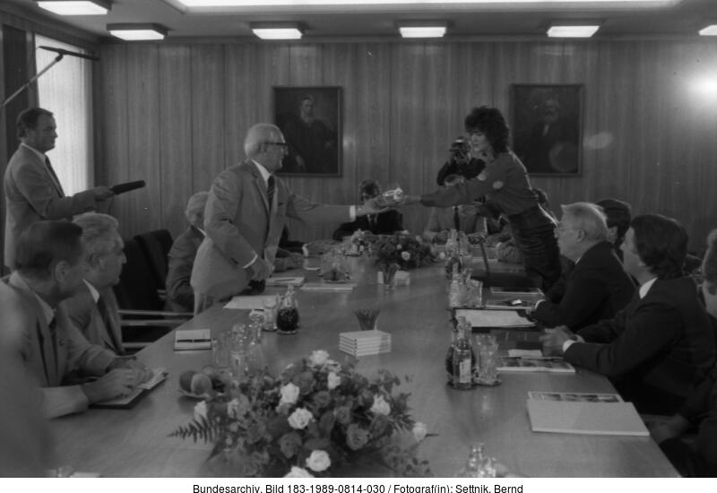 Eine Gruppe von Personen sitzt um einen Konferenztisch, während zwei Personen in der Mitte des Raums Dokumente austauschen; ein Fotograf hält den Moment fest.
