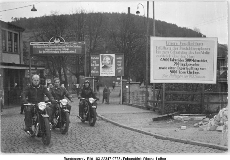 Drei Männer fahren auf Motorrädern auf einer Kopfsteinpflasterstraße in der Nähe großer Propagandaplakate und einer Toreinfahrt, mit Bäumen und Hügeln im Hintergrund.