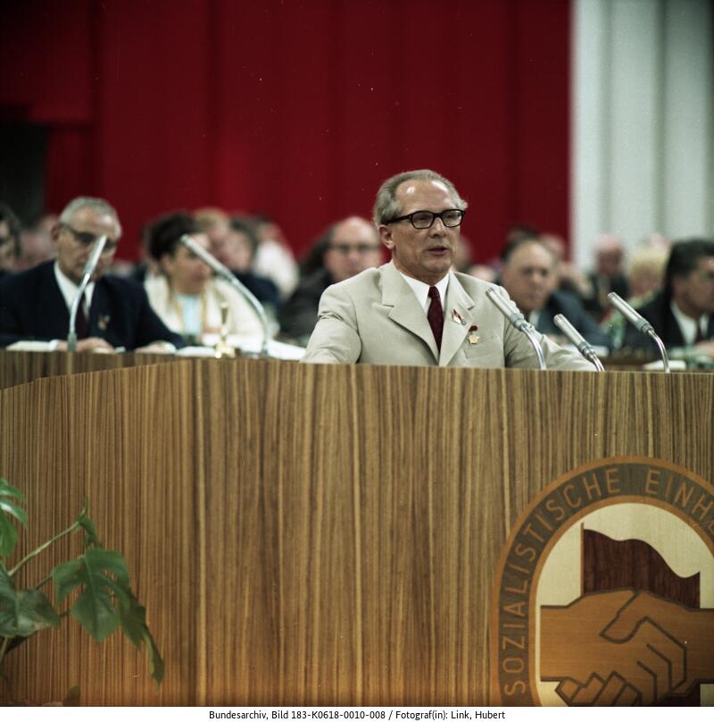 Ein Mann in einem beigen Anzug spricht während einer formellen Veranstaltung an einem Podium mit Mikrofonen, hinter dem mehrere Personen sitzen. Auf dem Podium ist ein Logo mit zwei ineinander verschränkten Händen zu sehen.