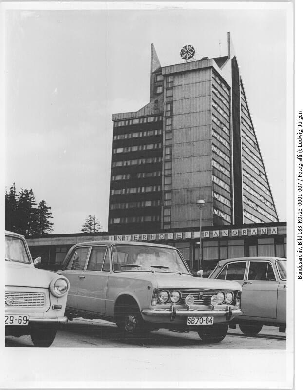Drei Autos parken vor dem Interhotel Panorama, einem hohen, modernistischen Gebäude mit kantigem Design und einem Wappen auf der Spitze.