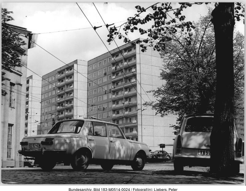 Zwei Autos sind auf einer Straße vor hohen, einheitlichen Wohnhäusern geparkt, im Vordergrund sind Bäume und Stromleitungen zu sehen.