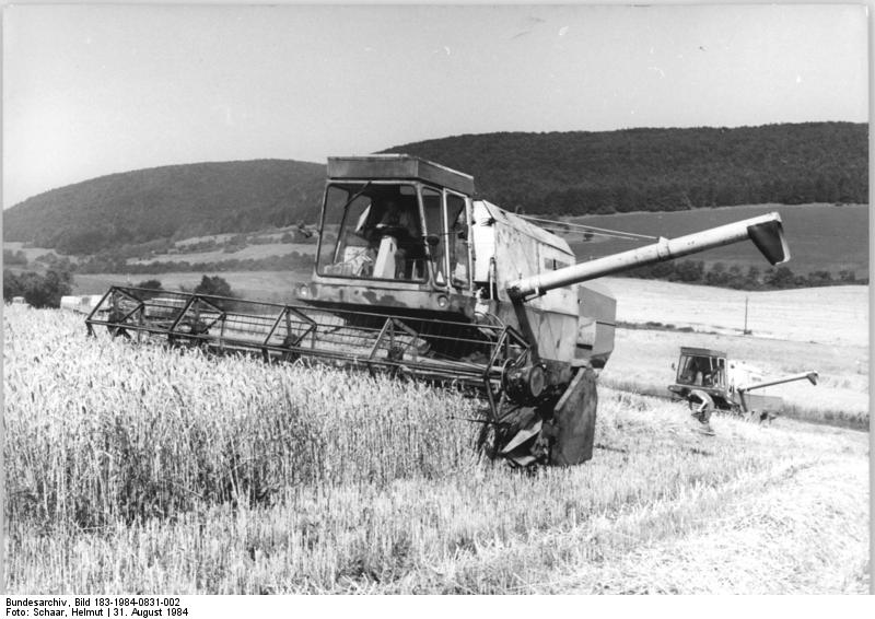Zwei Mähdrescher arbeiten tagsüber auf einem Getreidefeld mit Hügeln und Bäumen im Hintergrund.