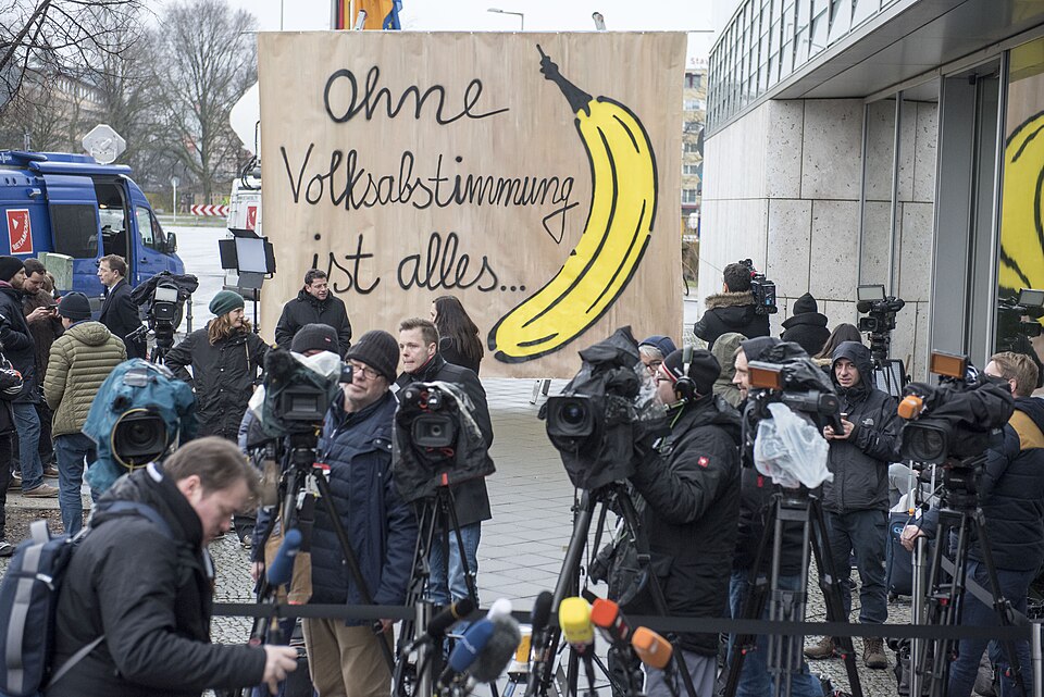 Eine Gruppe von Journalisten mit Kameras versammelt sich vor einem großen Schild mit einer Banane und dem deutschen Text "Ohne Volksabstimmung ist alles ..." vor einem modernen Gebäude.