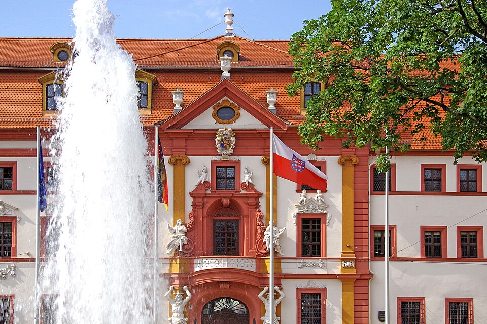 Ein großer Brunnen vor einem historischen Gebäude mit rotem Dach, verzierter Fassade und mehreren Fahnen, darunter eine mit einem Wappen.