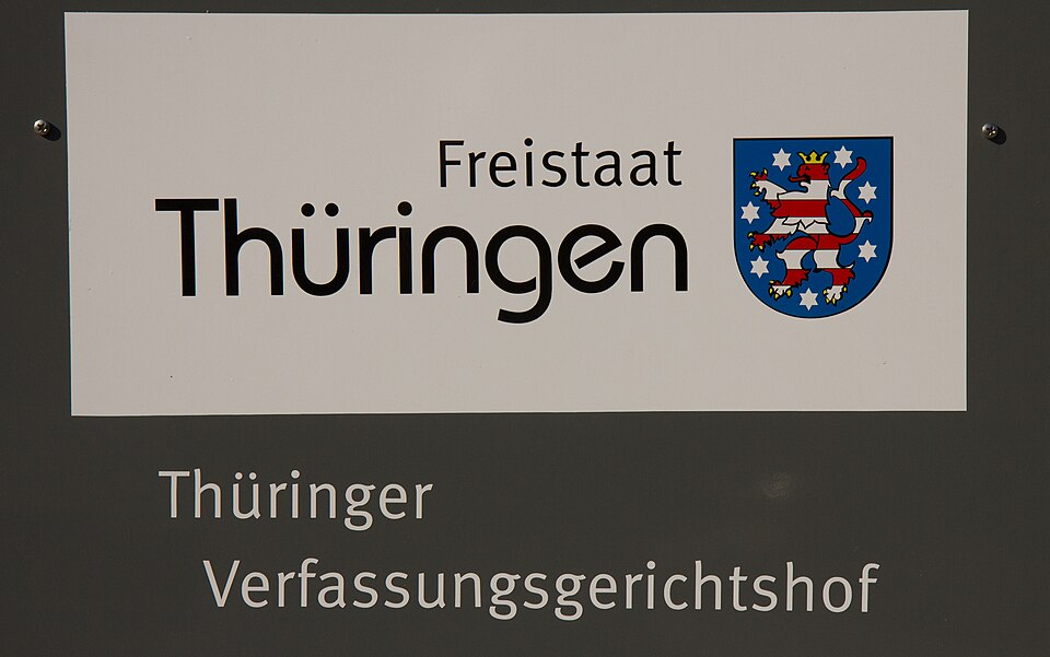 Schild mit der Aufschrift "Freistaat Thüringen" mit dem Thüringer Landeswappen über "Thüringer Verfassungsgerichtshof" auf grauem Grund.