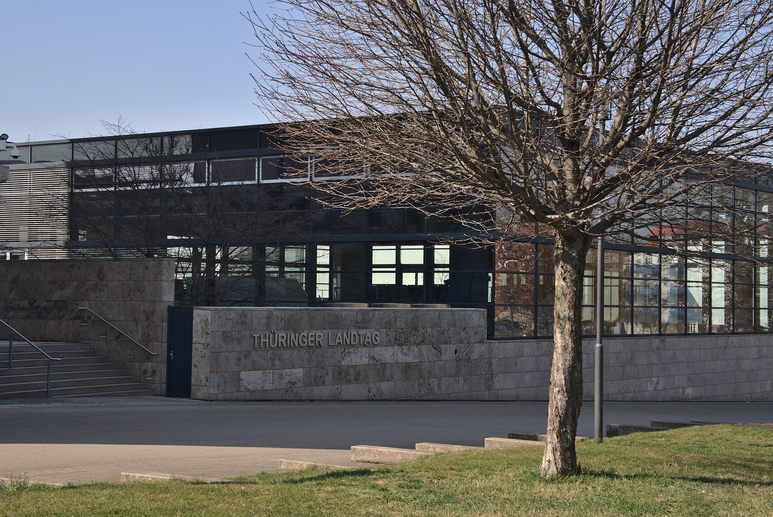 Ein modernes Gebäude aus Stein und Glas mit dem Schild "Thüringer Landtag" an der Wand, teilweise verdeckt durch einen blattlosen Baum im Vordergrund.