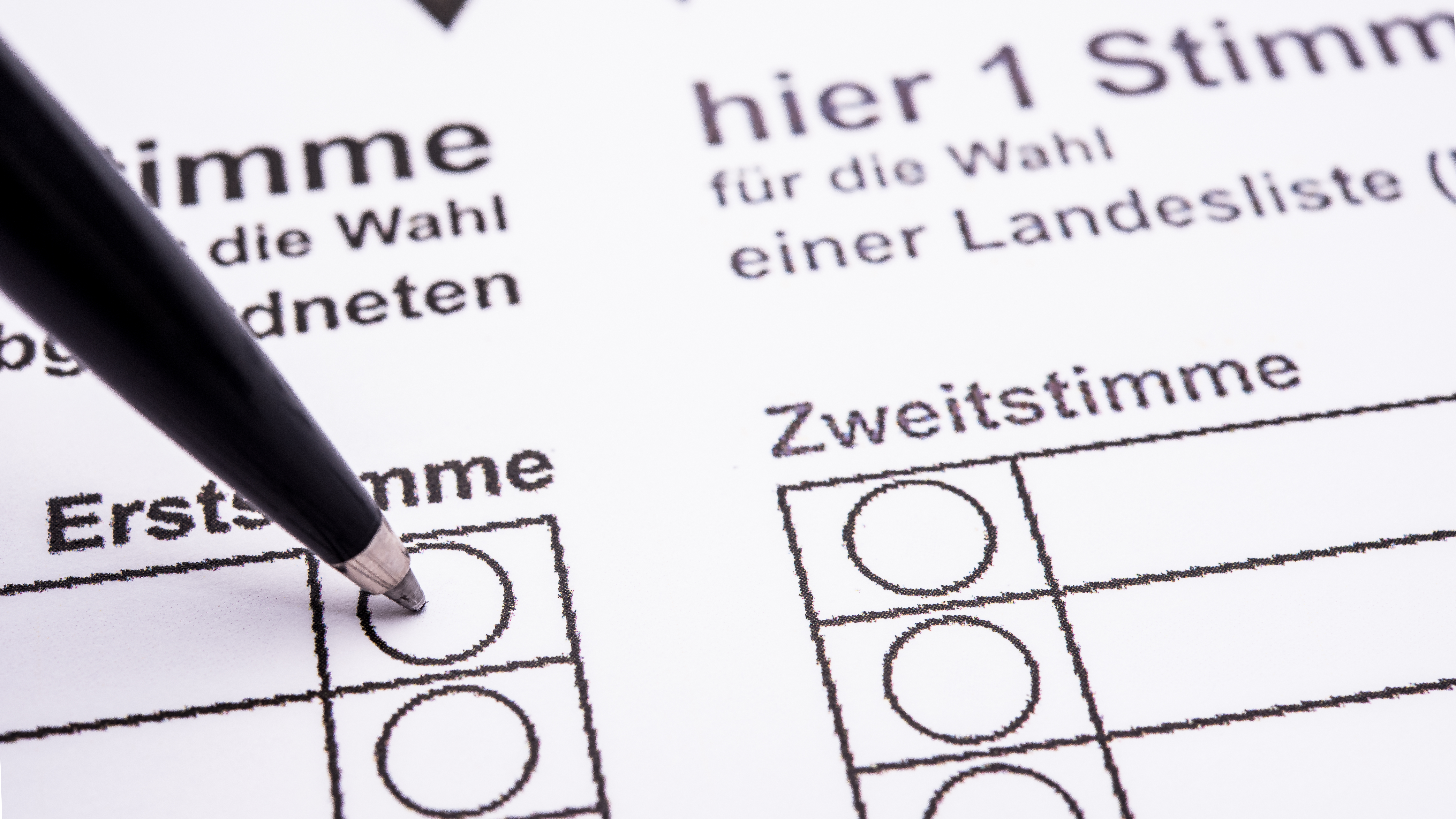 Ein schwarzer Stift markiert einen Kreis auf einem deutschen Wahlzettel unter "Erststimme", wobei Kästchen und Text auf dem Formular sichtbar sind.