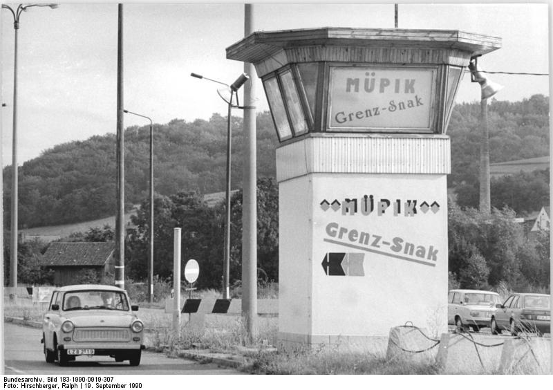 Ein Schwarz-Weiß-Foto zeigt einen kleinen Wachturm mit der Aufschrift "MÜPIK Grenz-Snack" am Rande einer Straße, mit einem herannahenden Auto und Hügeln im Hintergrund.