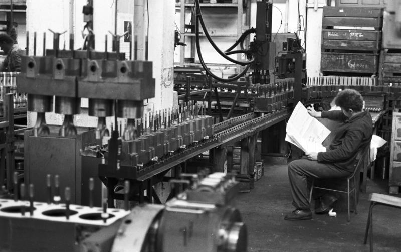 Eine Person sitzt auf einem Stuhl und liest eine Zeitung neben einem Fließband in einer Fabrik mit industriellen Maschinen und Anlagen.