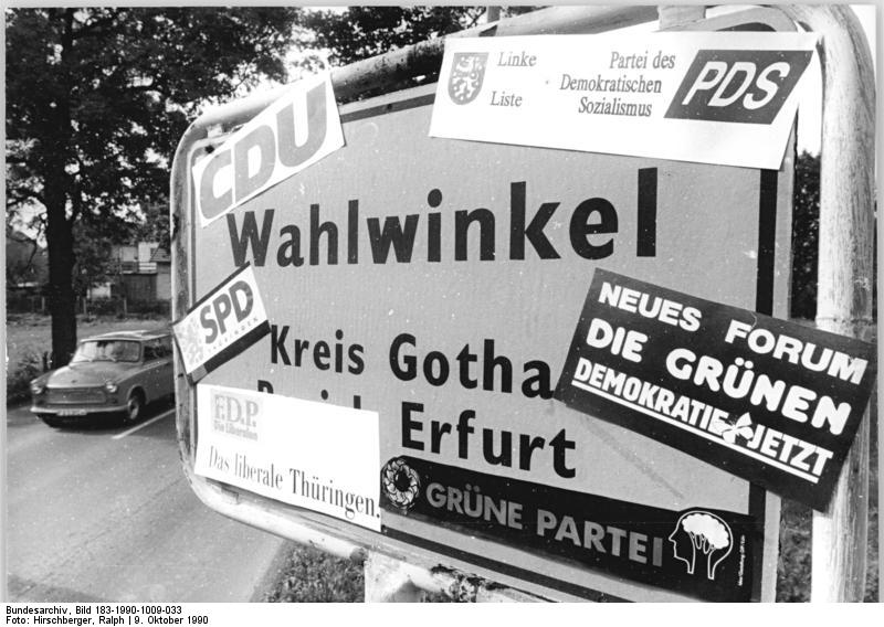 Ein Straßenschild mit Aufklebern verschiedener politischer Parteien, darunter CDU, SPD, PDS und Die Grünen.