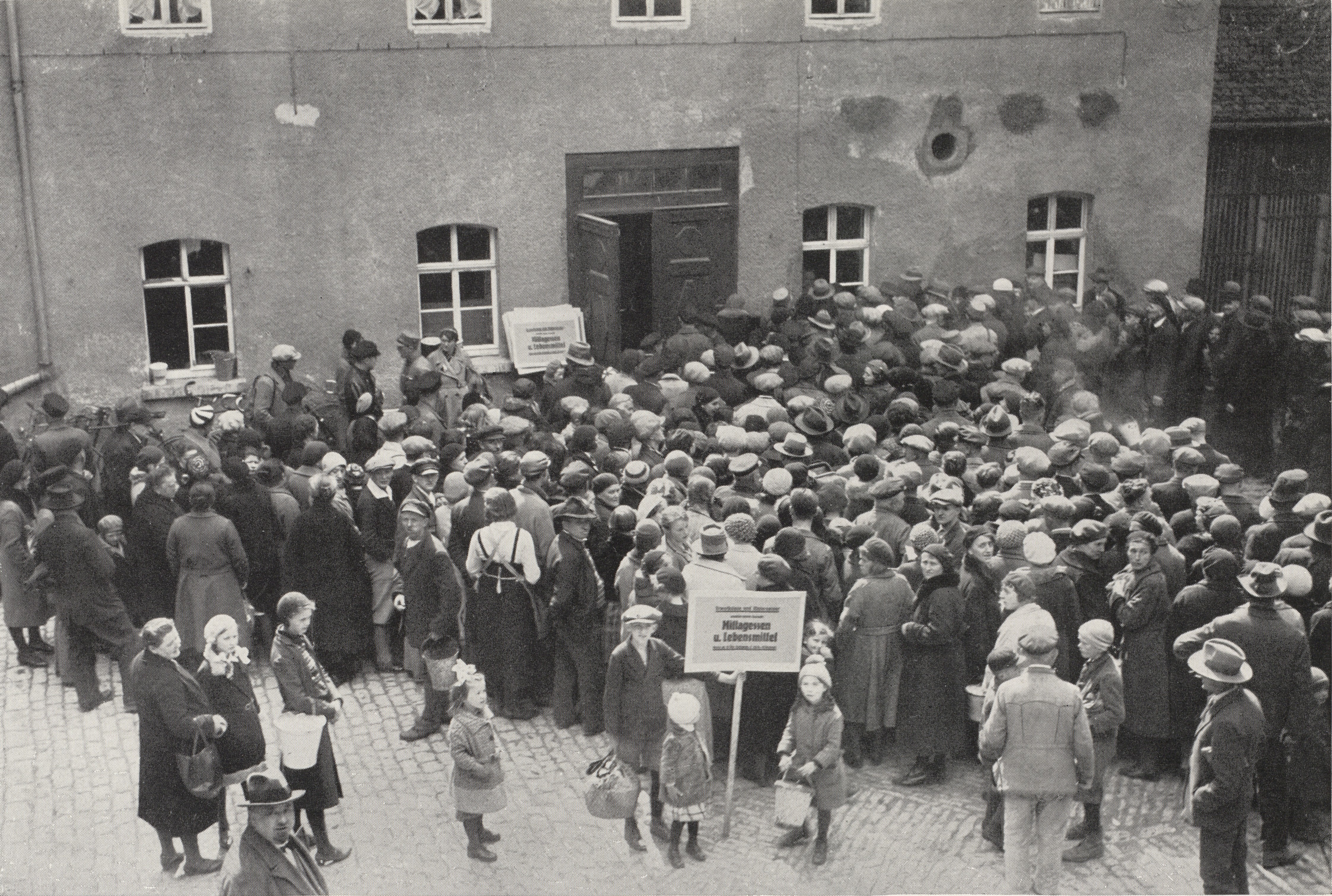 Eine große Menschenmenge, viele mit Hüten und Mänteln, versammelt sich vor einem mit Fenstern gesäumten Gebäude; einige halten Schilder, andere stehen Schlange.