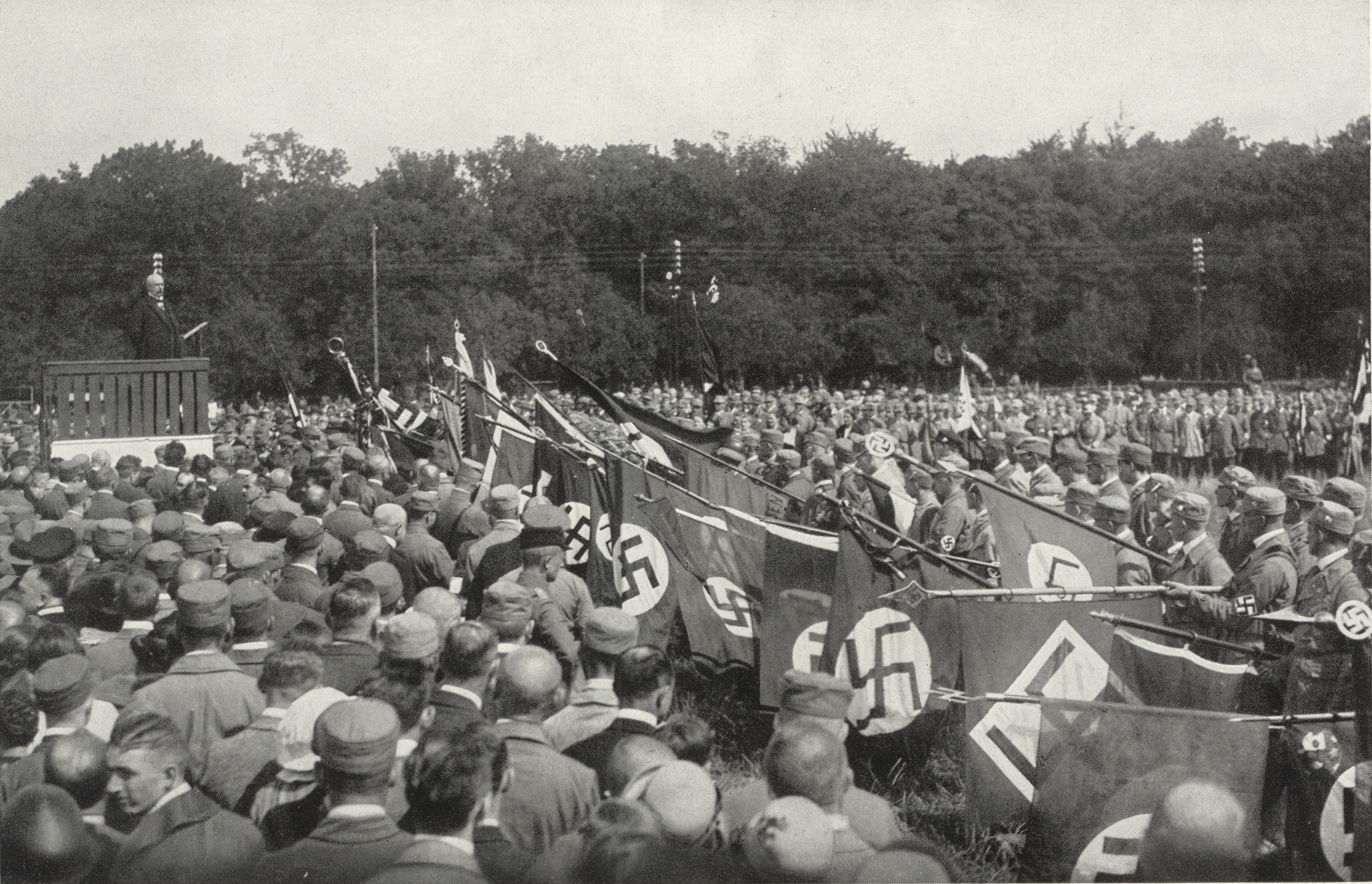 Große Menschenmenge bei einer Nazikundgebung in den 1930er Jahren mit Hakenkreuzfahnen, Uniformierten und einem Redner, der von einer Plattform im Freien zu den Zuhörern spricht.