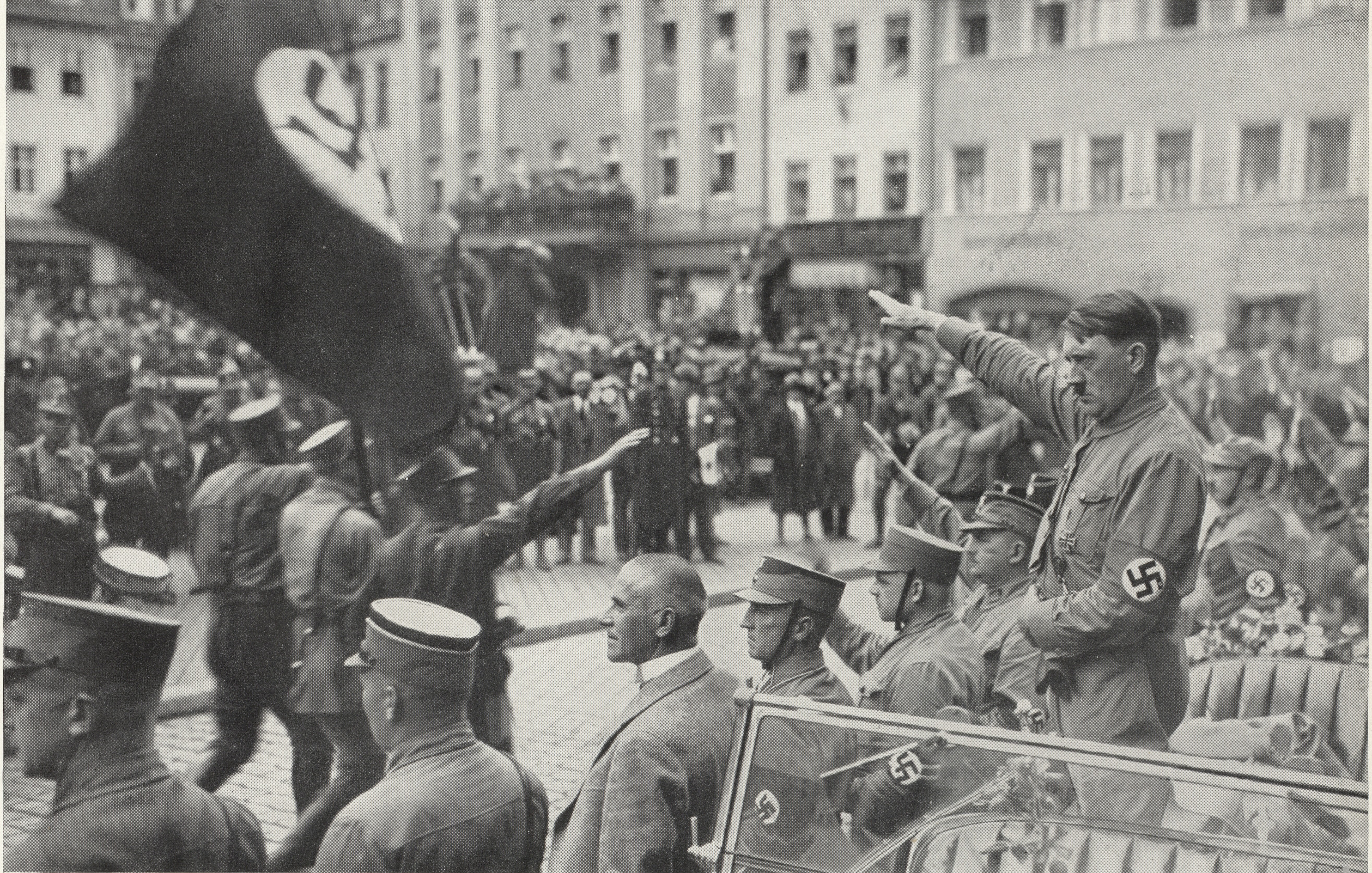 Ein historisches Schwarz-Weiß-Foto, das Adolf Hitler und uniformierte Beamte in einem Auto während einer Nazi-Parade zeigt, mit Fahnen und Menschenmengen im Hintergrund.