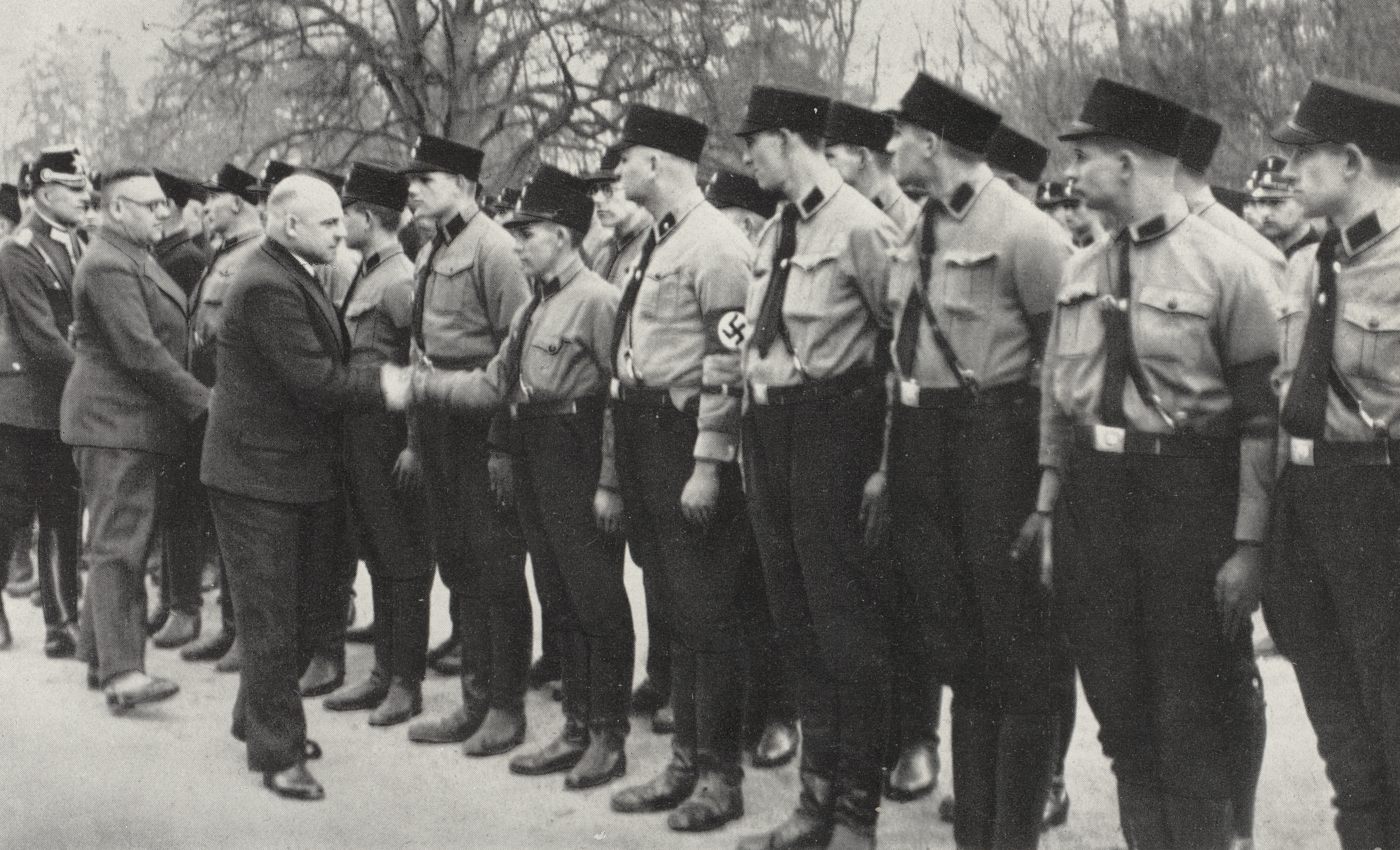 Eine Gruppe von Männern in Uniform, darunter Nazi-Armbinden, steht in einer Reihe im Freien; ein Mann im Anzug schüttelt einem von ihnen die Hand.