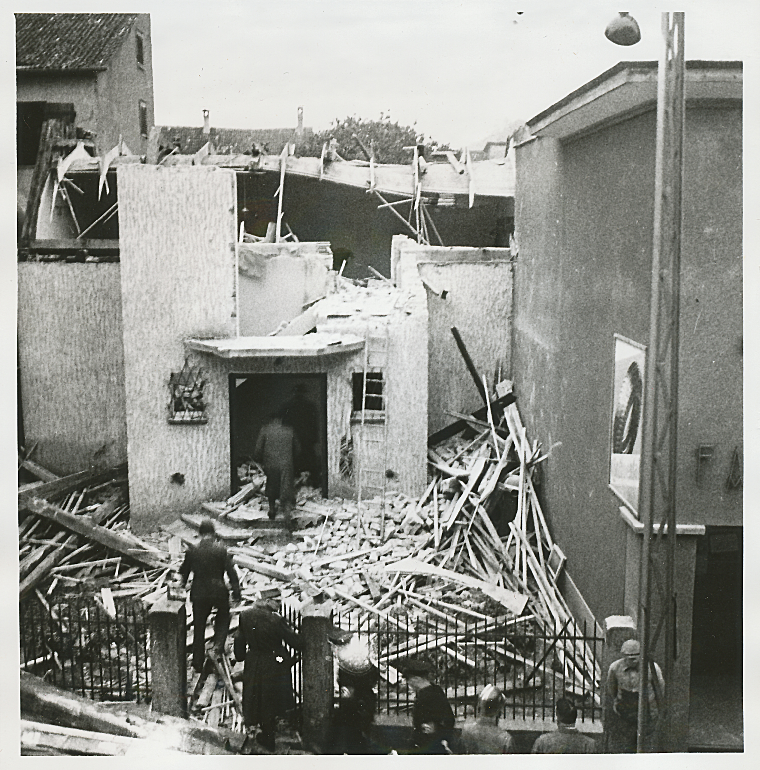 Eine Synagoge mit erheblichen strukturellen Schäden und Trümmern davor; mehrere Personen inspizieren den Schauplatz und laufen zwischen den Trümmern umher.
