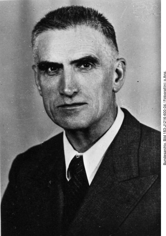 Schwarz-Weiß-Porträt eines älteren Mannes mit kurzen Haaren, der Anzug und Krawatte trägt und mit neutralem Gesichtsausdruck in die Kamera schaut.