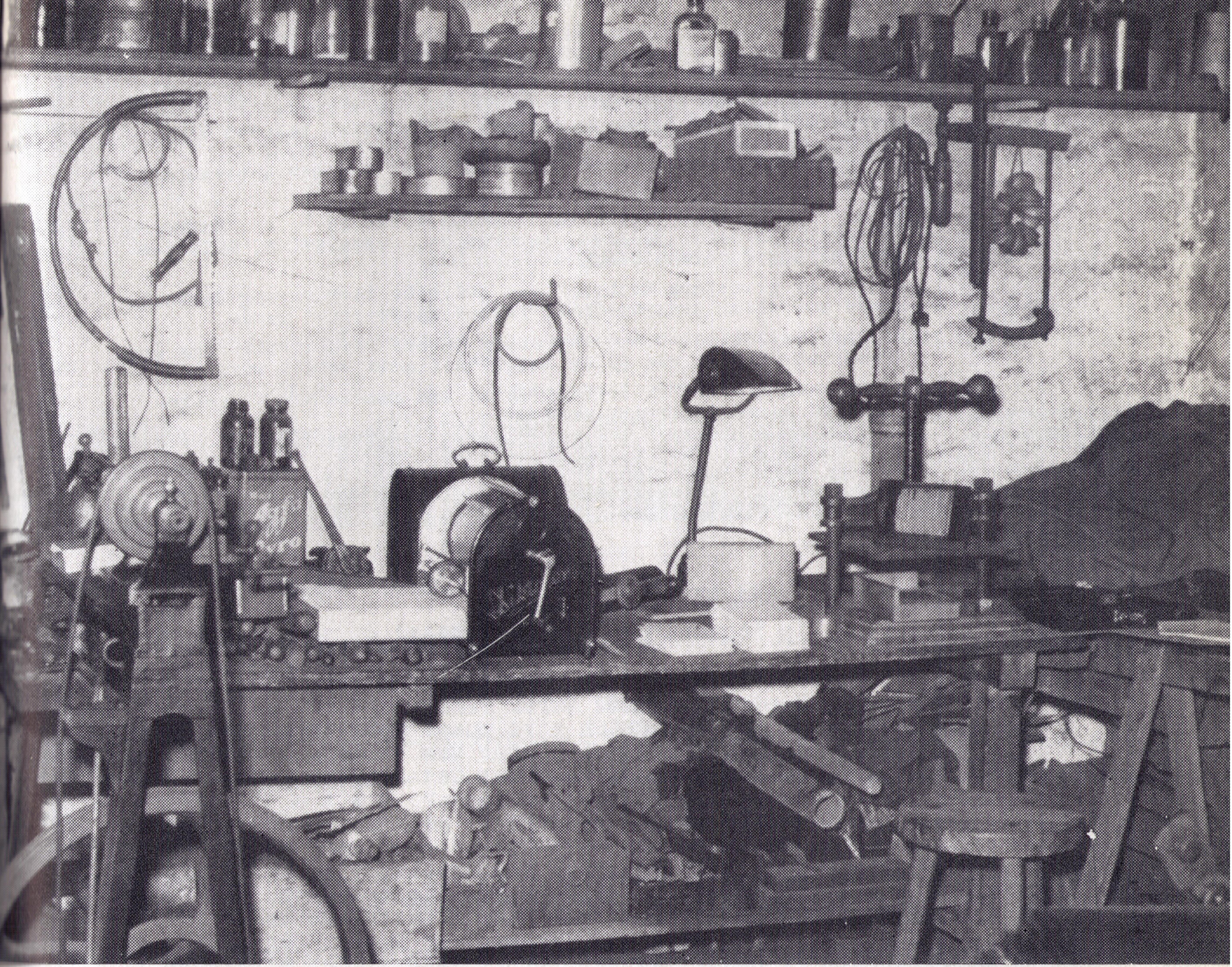 Eine überfüllte Werkstatt mit verschiedenen Werkzeugen, Maschinen, Regalen mit Gläsern und Dosen, einer Werkbank, einem Hocker und verstreuten Materialien.