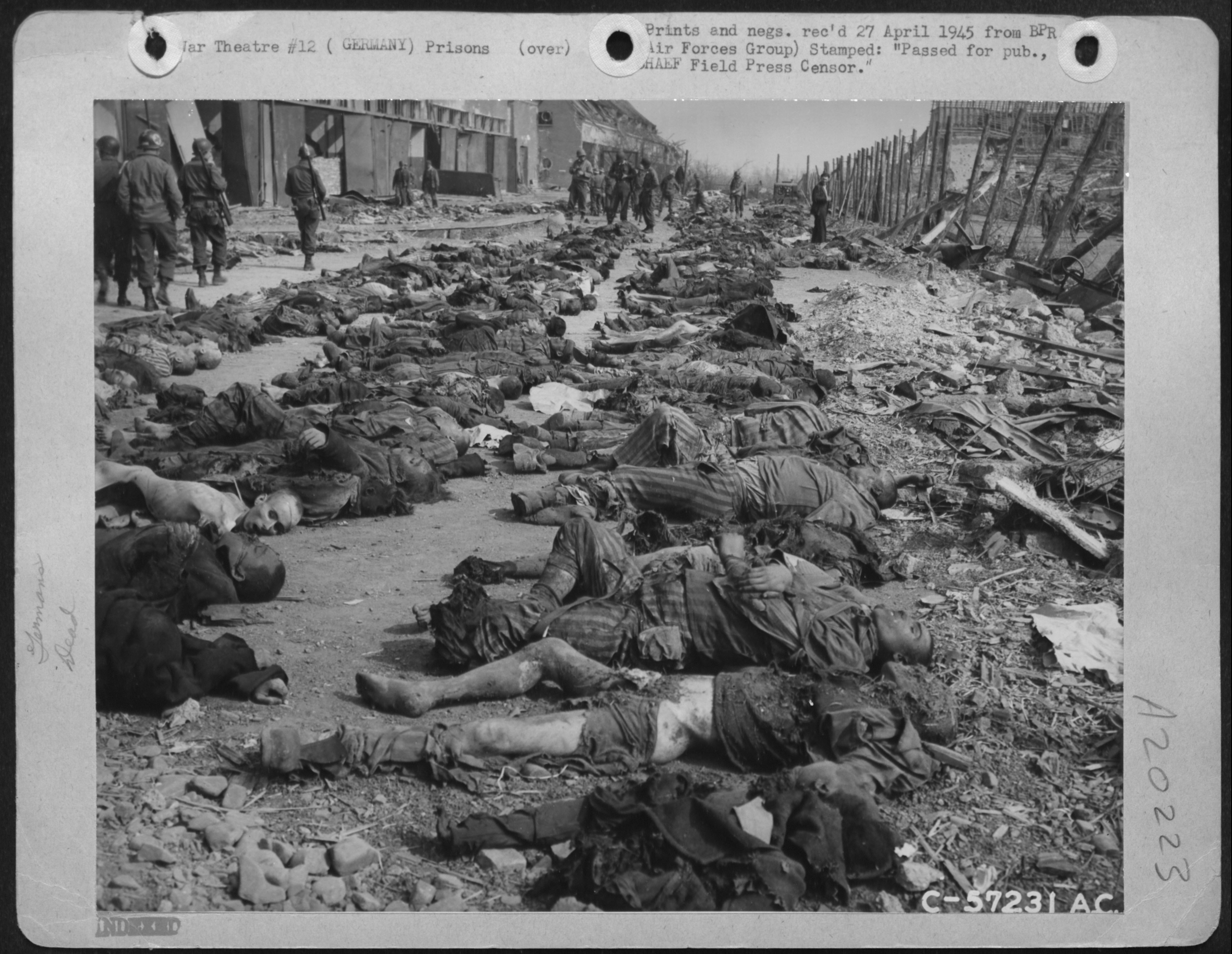 Schwarz-Weiß-Foto, das zahlreiche Leichen verstorbener Häftlinge zeigt, die in einem Konzentrationslager auf dem Boden liegen. Im Hintergrund sind Soldaten zu sehen, 1945.