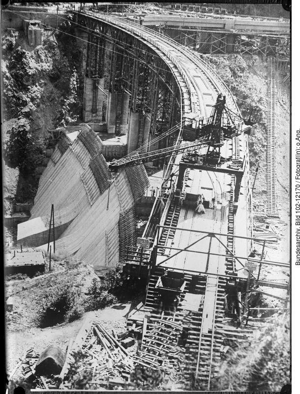 Historisches Schwarz-Weiß-Foto, das den Bau einer großen Eisenbahnbogenbrücke mit Arbeitern und Gerüsten über einer felsigen Landschaft zeigt.