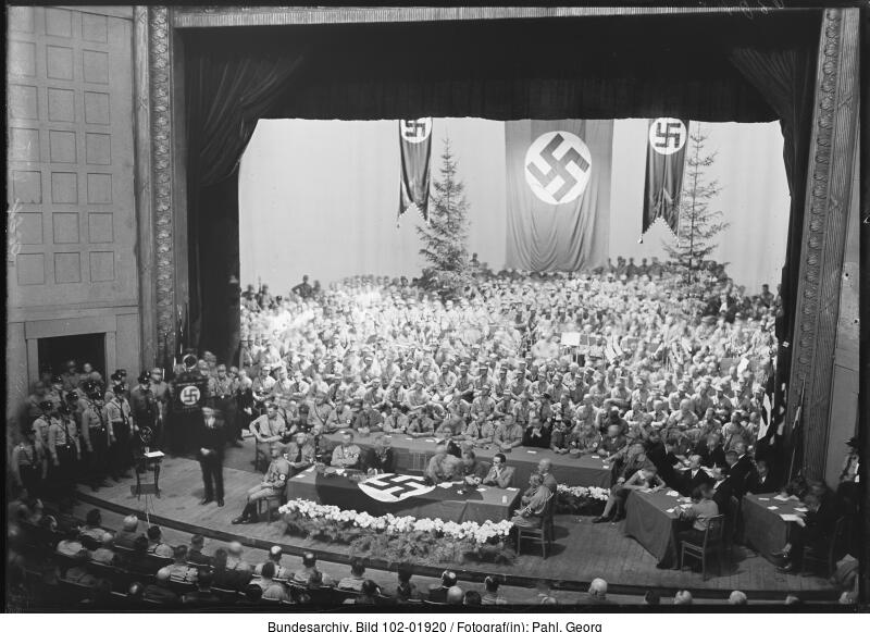 Eine große Versammlung in einem Theater mit Nazi-Fahnen und -Symbolen, mit uniformierten Beamten, die an Tischen auf der Bühne sitzen, und einem großen Publikum im Hintergrund.