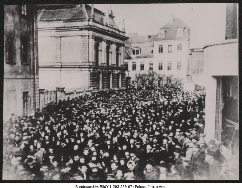 Eine große Menschenmenge versammelt sich vor mehreren mehrstöckigen Gebäuden in einer Stadt; die meisten tragen Mäntel und Mützen, was auf kaltes Wetter hindeutet.