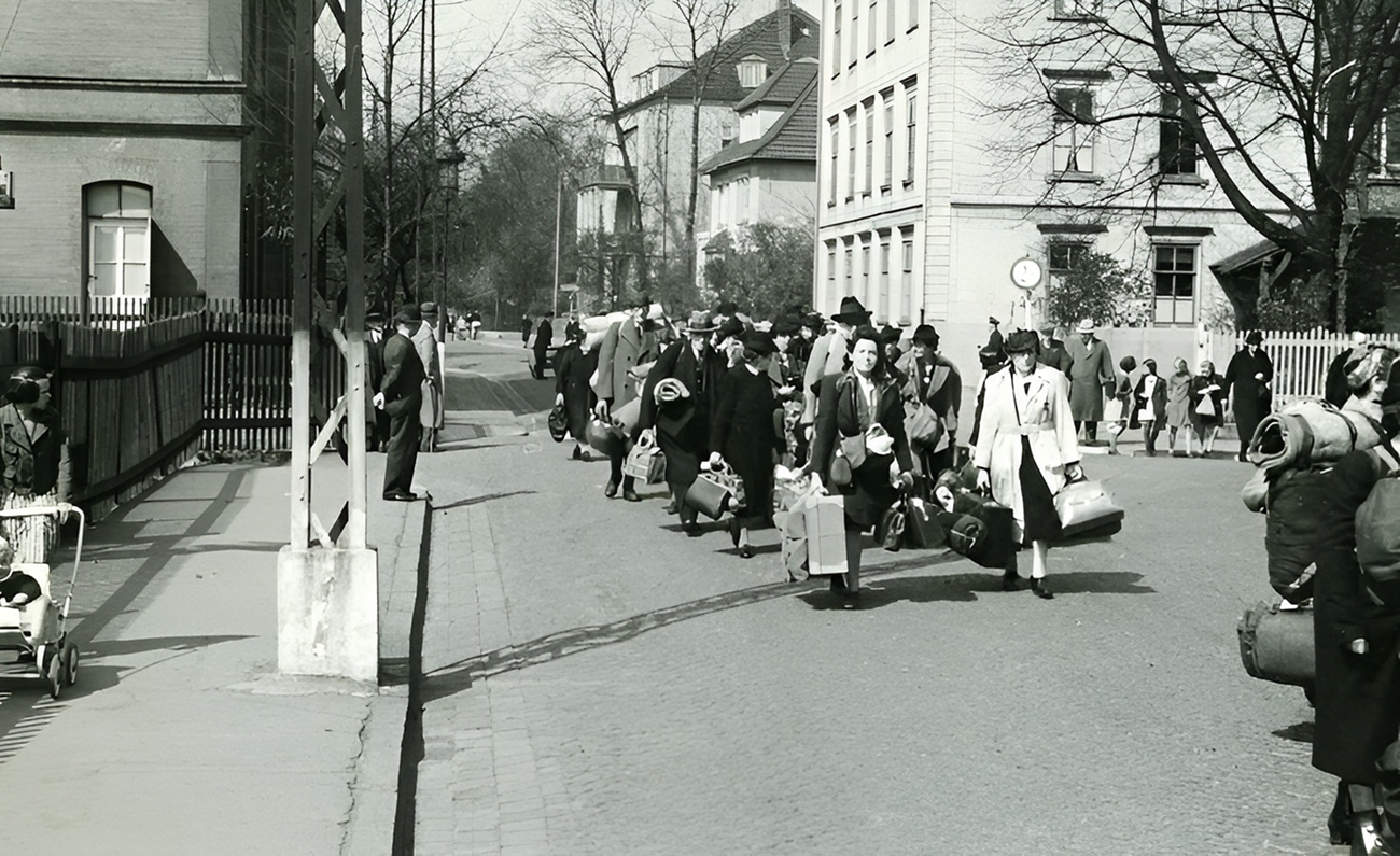 Eine Gruppe von Personen, die Gepäck und Taschen tragen, geht eine Straße entlang, während andere in der Nähe stehen oder beobachten. Die Szene scheint aus einer vergangenen Zeit zu stammen.