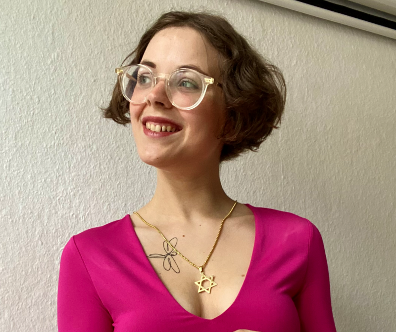 Eine Frau mit kurzen braunen Haaren und Brille lächelt, sie trägt ein rosafarbenes Oberteil, eine Halskette mit einem Davidstern und ein Blumentattoo auf der Brust.