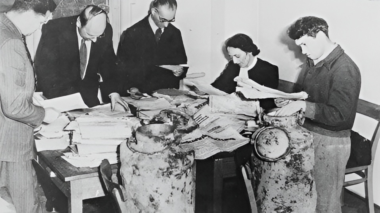 Fünf Personen untersuchen alte Manuskripte und Schriftrollen an einem Tisch, in dessen Nähe mehrere große, verwitterte Krüge stehen.