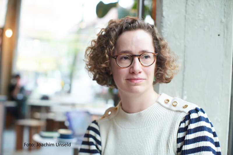 Eine Frau mit lockigem Haar und Brille, die einen gestreiften Pullover trägt, sitzt in einem Innenraum an einem Fenster und schaut leicht zur Seite. Bildnachweis: Joachim Unseld.