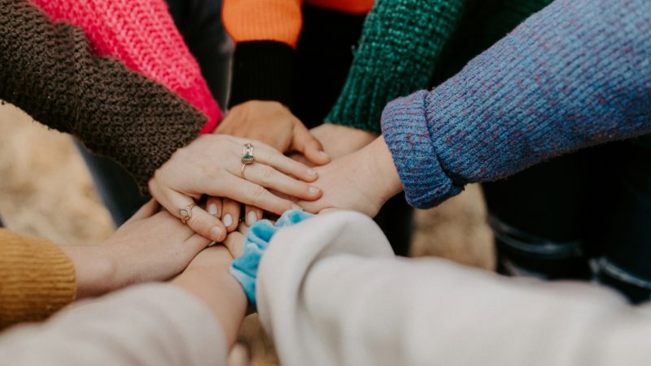 Eine Gruppe von Menschen steht im Kreis und jeder legt eine Hand in die Mitte, um Teamwork und Einigkeit zu zeigen.