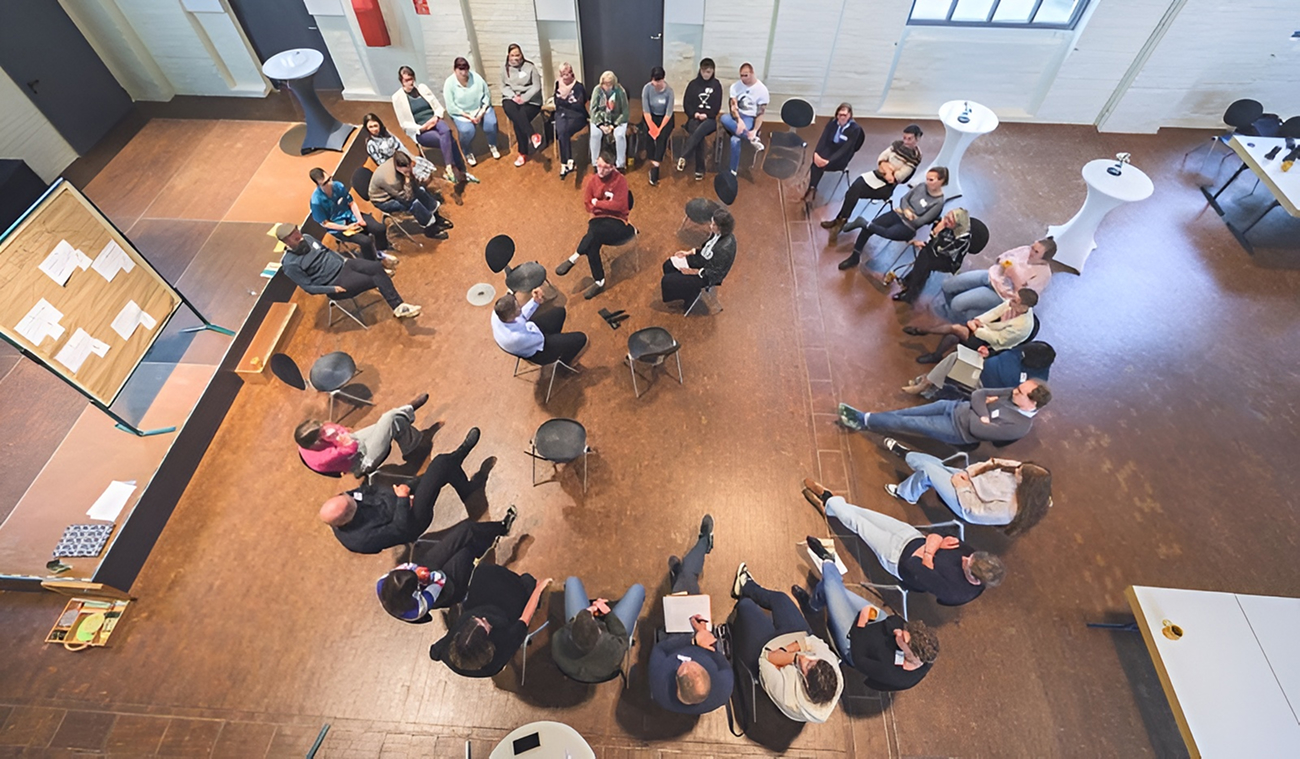 Eine Gruppe von Menschen sitzt in einem großen Kreis auf Stühlen in einem geräumigen Raum und führt eine Diskussion oder ein Treffen durch.