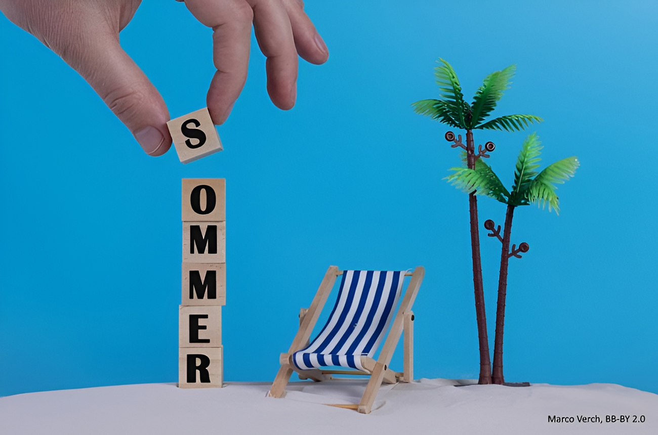 Eine Hand stapelt Holzklötze mit der Aufschrift "SOMMER" neben einem gestreiften Liegestuhl und einer kleinen Plastikpalme auf einer sandigen Fläche mit blauem Hintergrund.